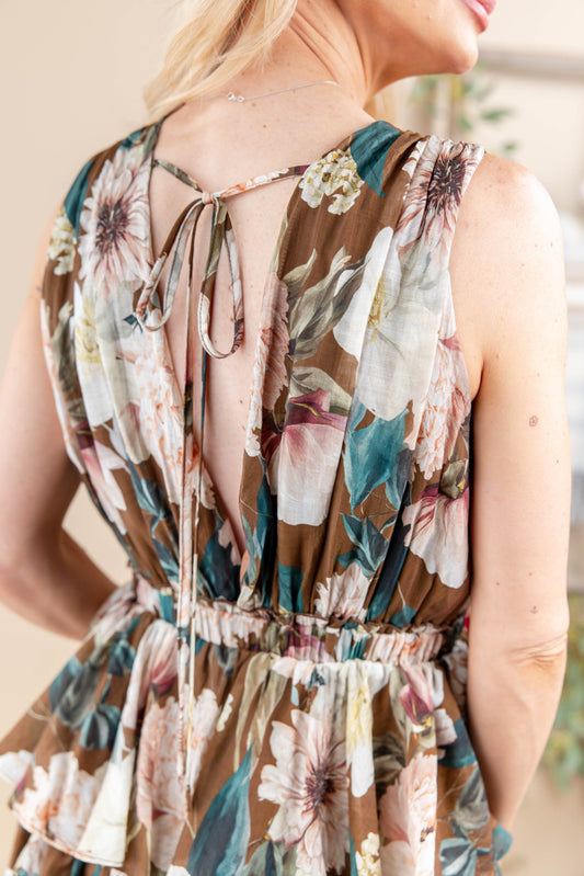 Close-up back view of a woman wearing a sleeveless floral dress with a tie-back neckline and gathered waist, shown in a softly lit indoor setting.