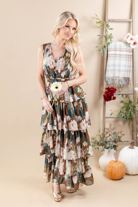 A woman models a sleeveless, tiered maxi dress with a floral print in earthy tones of brown, beige, and teal. She is standing in a styled studio setting with a wooden ladder, a plaid throw blanket, and decorative pumpkins and vases.