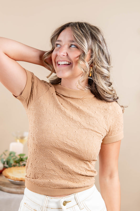 A woman wearing a beige textured short-sleeve mock neck top and white pants, smiling and looking upward in a warm indoor setting.