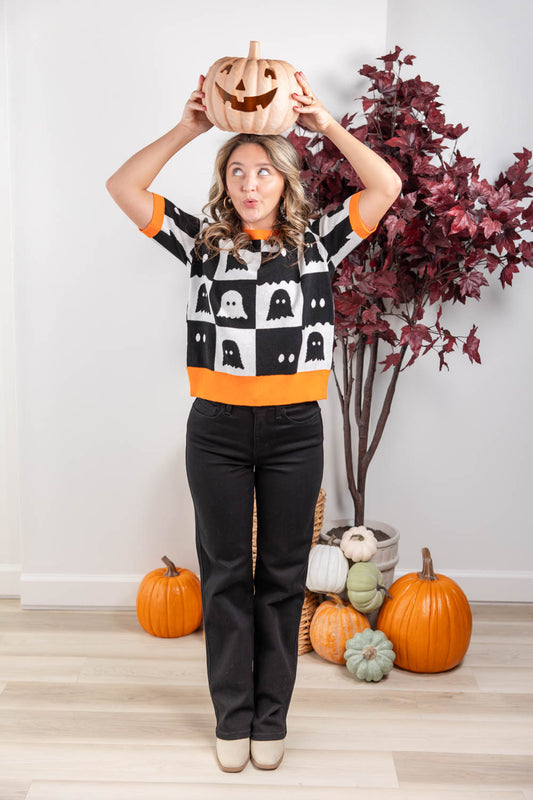 A woman wearing a black and white Halloween sweater with ghost patterns and orange trim, holding a carved pumpkin above her head. She is standing in a decorated room with pumpkins and a potted plant.
