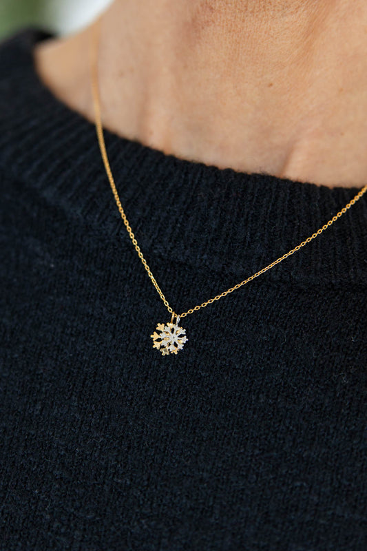 A close-up of a person wearing a delicate gold chain necklace with a small snowflake-shaped pendant set with clear stones, paired with a black knit sweater.