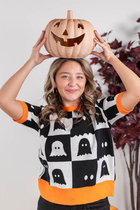 A woman wearing a black and white checkered sweater with orange trim and ghost patterns, holding a carved jack-o'-lantern pumpkin above her head against a light gray background with a decorative plant.