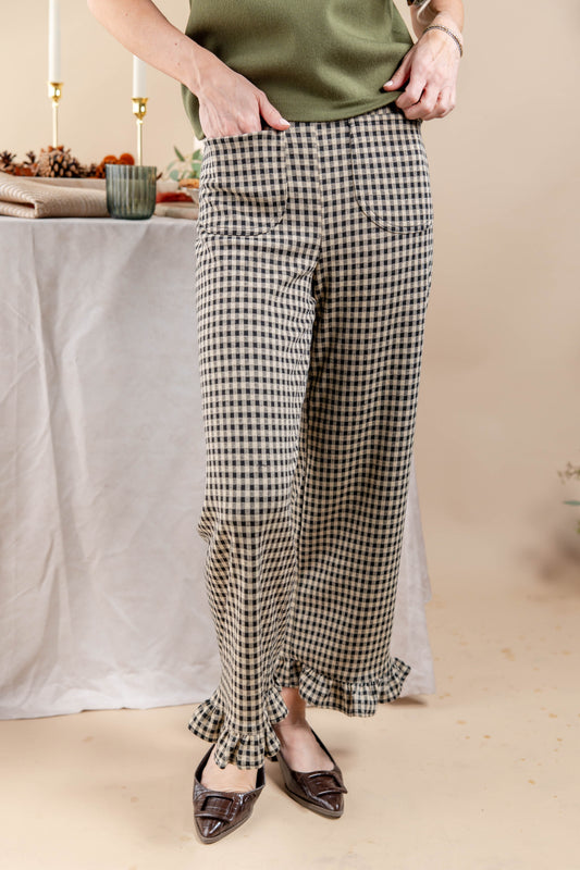 A woman wearing black and beige gingham wide-leg pants with ruffled hems, paired with a green top and brown pointed flats, standing in a styled indoor setting.