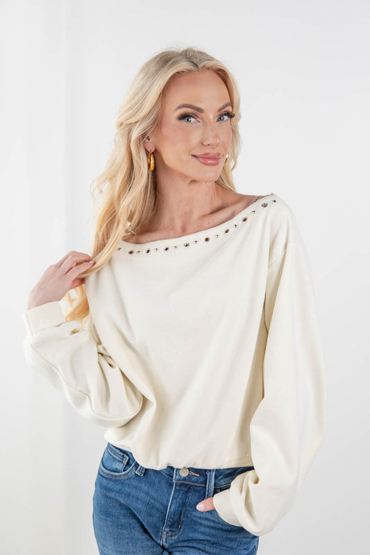 A blonde woman wearing a cream-colored, long-sleeve off-the-shoulder top with silver stud detailing along the neckline, paired with blue jeans, against a plain white background.