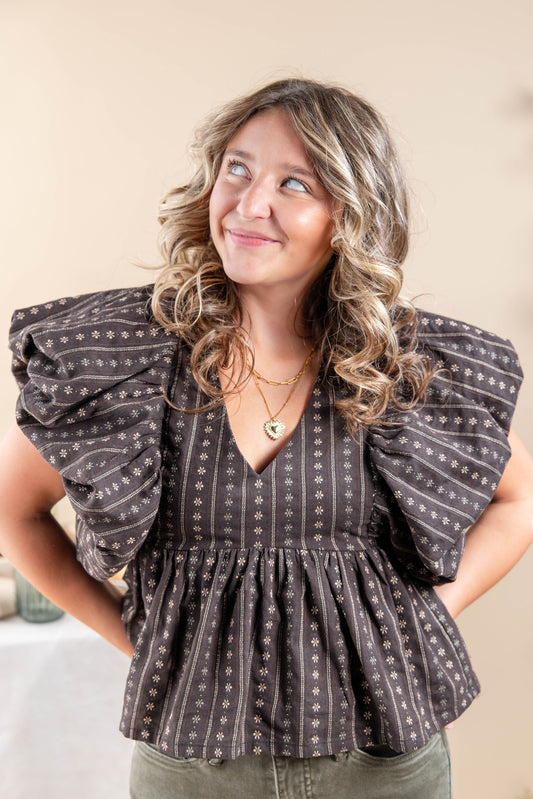 A woman wearing a dark brown and beige patterned puff-sleeve blouse, paired with olive green pants, posing against a neutral beige background.