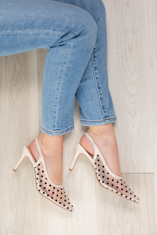 A person wearing light blue cropped jeans and sheer beige pointed-toe slingback heels with black polka dots, seated on a light wood floor.