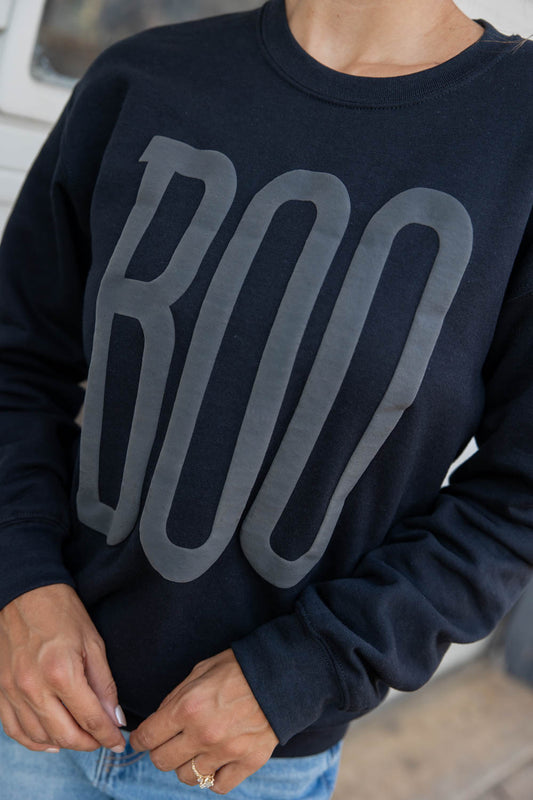 Close up of a black crewneck sweatshirt with a large gray 'BOO' graphic on the front, paired with light blue jeans