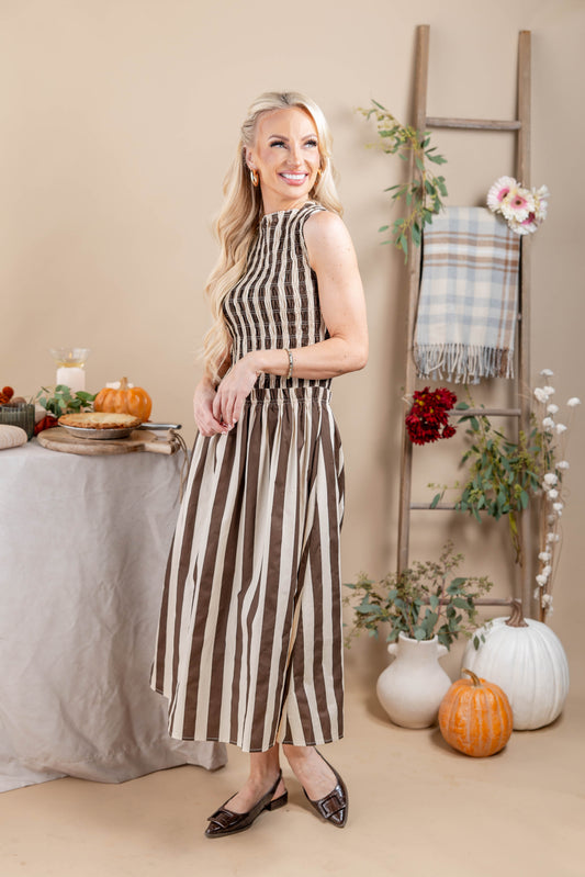 A woman modeling a sleeveless midi dress with vertical brown and beige stripes, standing in a styled indoor autumn setting with pumpkins, a table, and a ladder backdrop.