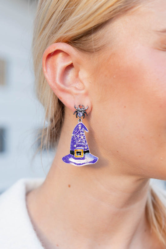 A close-up of a model's ear wearing a dangling earring shaped like a purple witch hat with a small spider charm at the top.