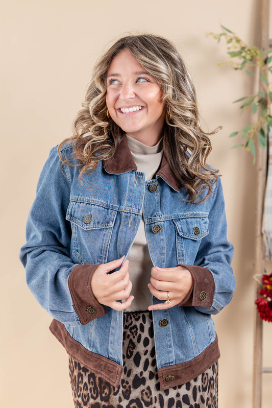 A woman wearing a blue denim jacket with brown suede-like collar, cuffs, and hem, layered over a beige top and leopard print skirt, posing against a warm beige background.