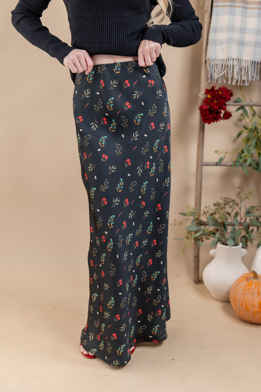 A woman wearing a long black maxi skirt with a small multicolor floral print, paired with a dark ribbed sweater. The photo is taken in a styled indoor setting with a beige backdrop, a wooden ladder, and autumn decorations like a pumpkin and potted plants.