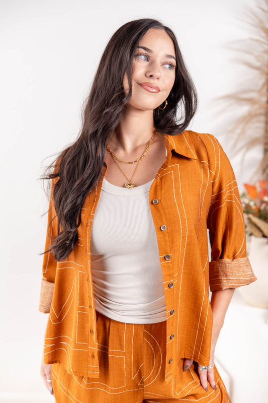 A woman wearing a matching burnt orange linen two-piece set with white line-art patterns, featuring a long-sleeve button-up shirt and coordinating pants, styled over a white ribbed tank top.