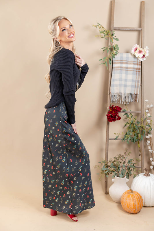 A woman posing in a black ribbed long-sleeve sweater and a black maxi skirt with a red and yellow floral pattern, standing in a studio decorated with a wooden ladder, pumpkins, and potted plants.