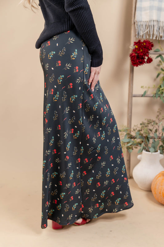 A woman wearing a long black maxi skirt with a colorful floral and leaf print, paired with a dark ribbed sweater. The photo is taken in a warm, neutral-toned indoor setting with decorative plants and a pumpkin in the background.