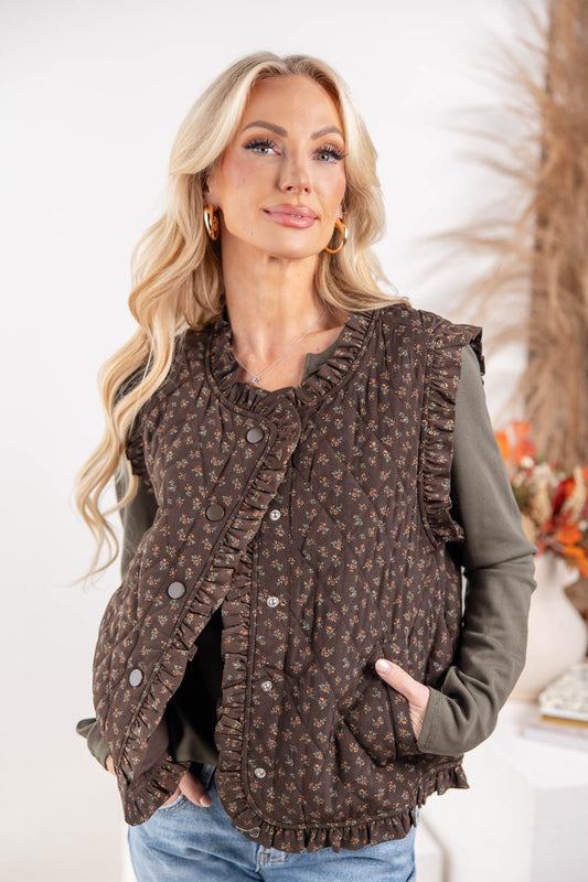 A woman wearing a brown ruffled button-front vest with a small floral pattern over a dark olive green long-sleeve shirt and blue jeans.