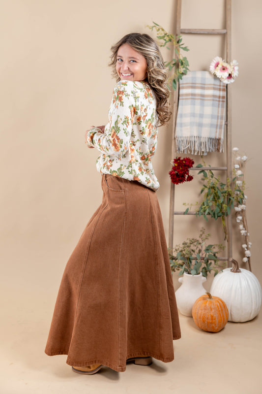 A woman posing in a studio setting, wearing a long-sleeve white floral shirt and a brown corduroy maxi skirt. She is standing next to a rustic wooden ladder decorated with a plaid throw blanket, flowers, and pumpkins against a solid beige background.