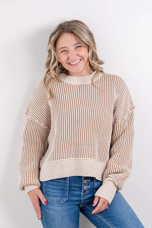 A smiling woman with blonde hair wearing a beige and cream vertically striped knit sweater and blue jeans, posing against a light gray studio background.