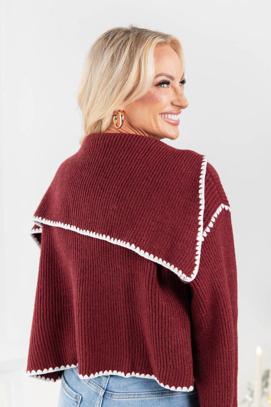 A woman wearing a burgundy ribbed knit cropped sweater with a wide triangular collar trimmed in white picot trim, shown from a three-quarters back view.