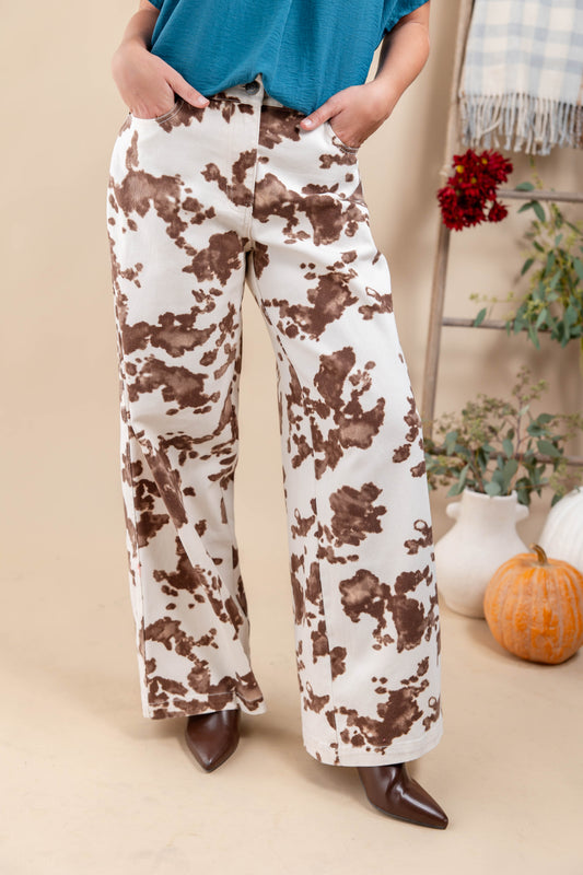 A woman wearing wide-leg pants with a brown and cream cow-print pattern, paired with a teal top and brown pointed-toe heels, standing in a styled indoor setting with pumpkins and a ladder.