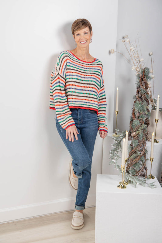 A woman wearing a long-sleeve knit sweater with horizontal stripes in red, green, orange, and blue, paired with blue jeans and beige slip-on shoes.