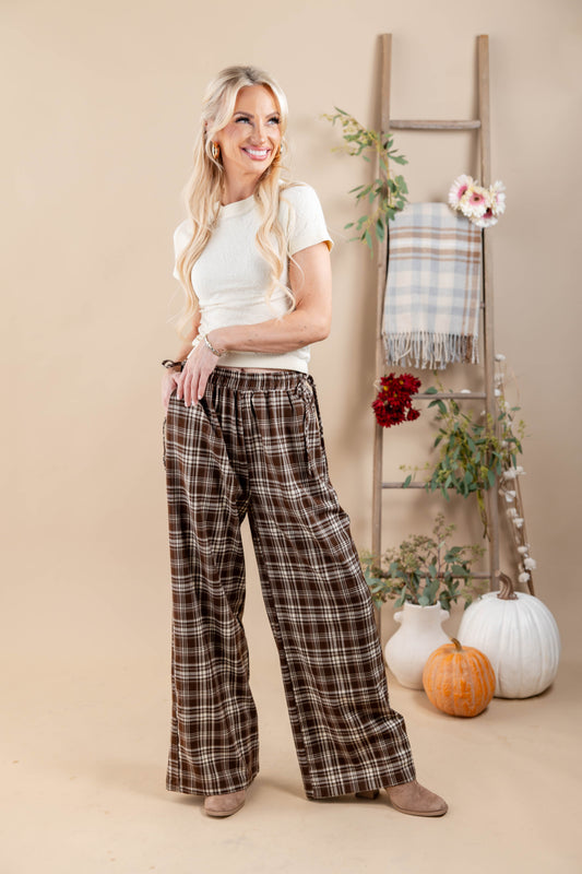 A woman stands in a studio wearing brown and beige plaid wide-leg pants with a white t-shirt. The background features a beige backdrop, a wooden ladder decorated with a plaid throw blanket and flowers, and two pumpkins on the floor.