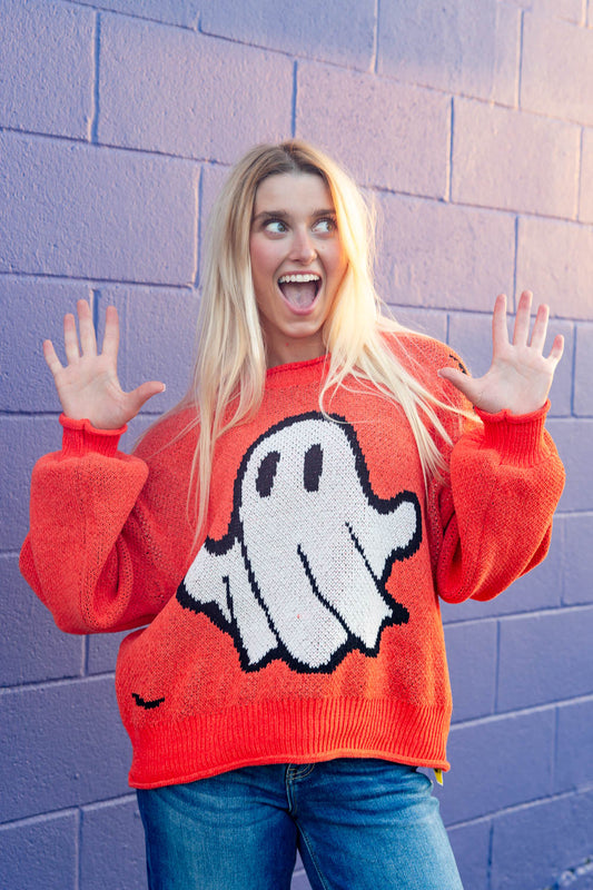 A smiling woman stands with hands up against a purple painted brick wall wearing a bright orange crewneck sweater with a large white ghost graphic.