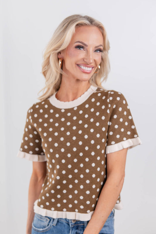 A woman wearing a brown short-sleeve knit top with white polka dots and cream ruffle trim at the collar, cuffs, and hem.