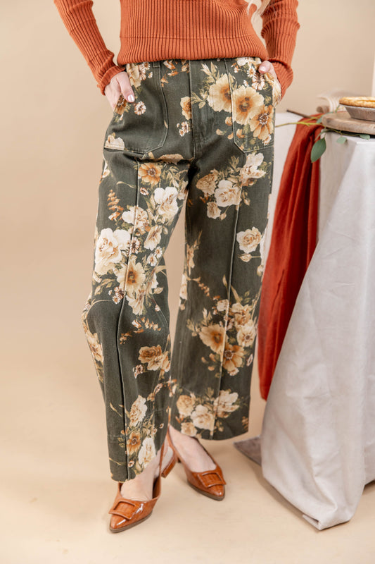 A person wearing olive green wide-leg pants with a beige and white floral print, styled with an orange ribbed sweater and brown pointed-toe heels, standing against a beige studio backdrop.