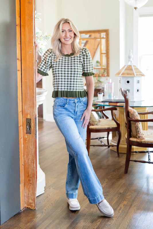 A woman stands in a bright, modern room wearing a green and white gingham short-sleeve top with dark green ruffle trim, paired with light-wash blue flare jeans and white sneakers.