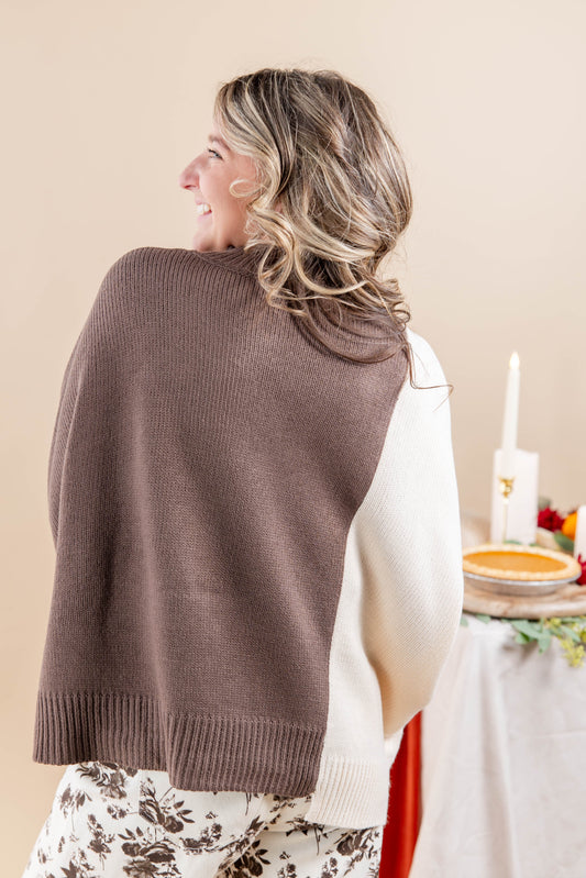 A woman shown from the back wearing a brown and cream two toned sweater, standing in a softly lit indoor setting with a table and candle in the background.