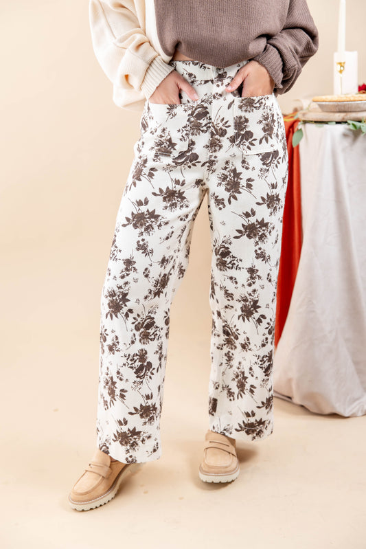 A woman wearing wide-leg white pants with a dark brown floral pattern, styled with a beige sweater and tan loafers, standing against a warm beige background.