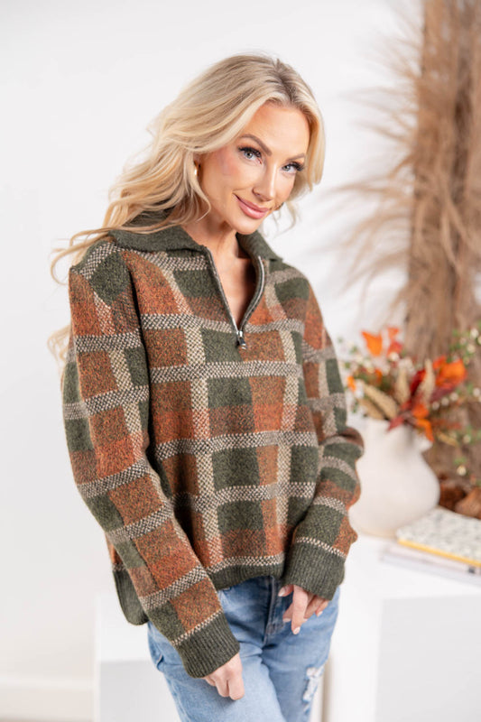 A woman wearing a long-sleeve quarter-zip knit sweater with a brown, green, and beige plaid pattern, paired with blue jeans.