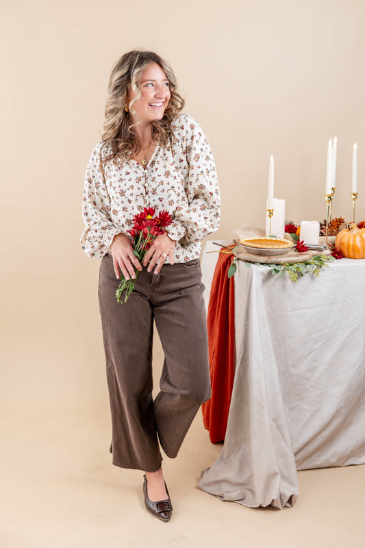 A woman stands in a studio setting wearing a cream blouse with a small floral print and brown wide-leg pants, holding a small bouquet of red flowers. To her right is a styled table with a gray tablecloth, candles, pumpkins, and autumn decor against a warm beige background.
