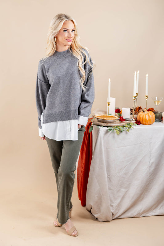 A woman models a gray knit sweater with a contrasting white hem, paired with olive green pants, standing next to a styled autumn table with pumpkins, candles, and a pie.