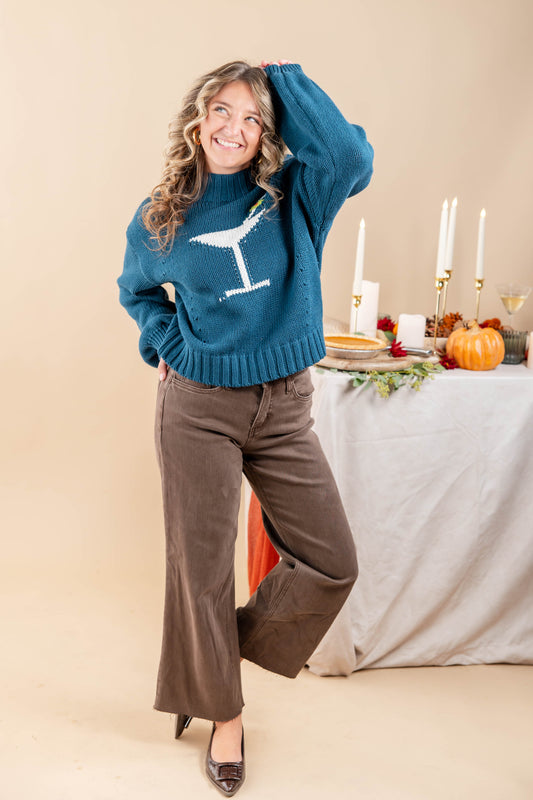 A woman models a teal knit sweater featuring a white martini glass graphic on the chest, paired with brown wide-leg pants and black heels, posed in front of a decorated table with pumpkins and candles.