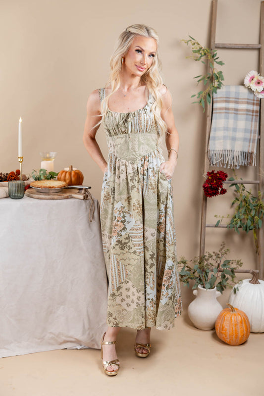 A woman models a sleeveless, maxi dress with a beige and green autumnal floral and landscape print. The dress features a smocked bodice with tie details at the shoulders and side pockets.