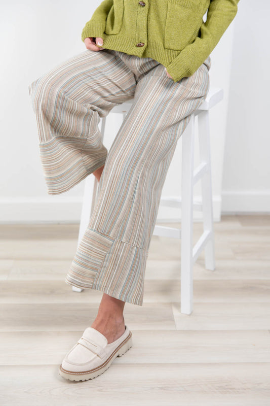 A person seated on a white stool wearing wide-leg striped pants in beige, cream, and muted blue tones, with a green cardigan and cream loafers in a bright indoor room.