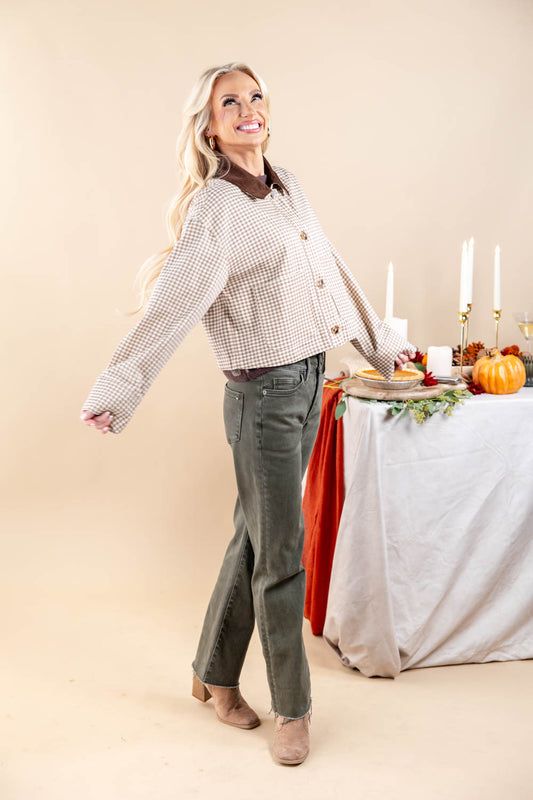 A woman modeling a cropped beige and white gingham button-up jacket with a brown collar, paired with olive green pants and tan boots, standing next to a styled autumn table.