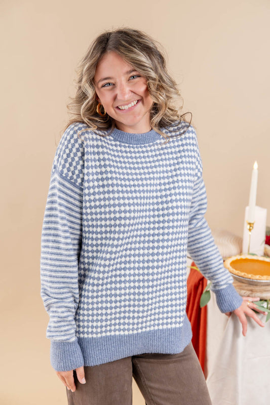 A smiling woman wearing a light blue and white checkerboard knit sweater, standing in a warm beige studio setting with a decorated table in the background.