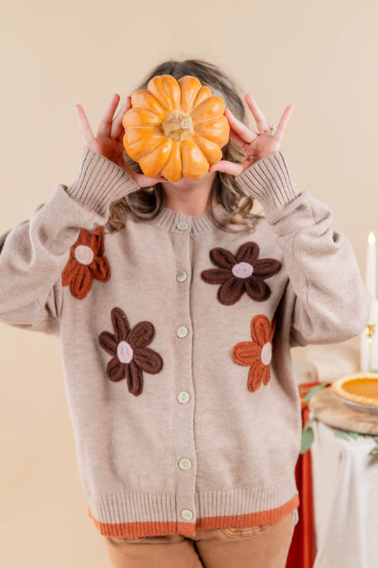 A person wearing a beige knit cardigan with brown and orange flower appliqués, holding a small orange pumpkin in front of their face against a warm beige background.