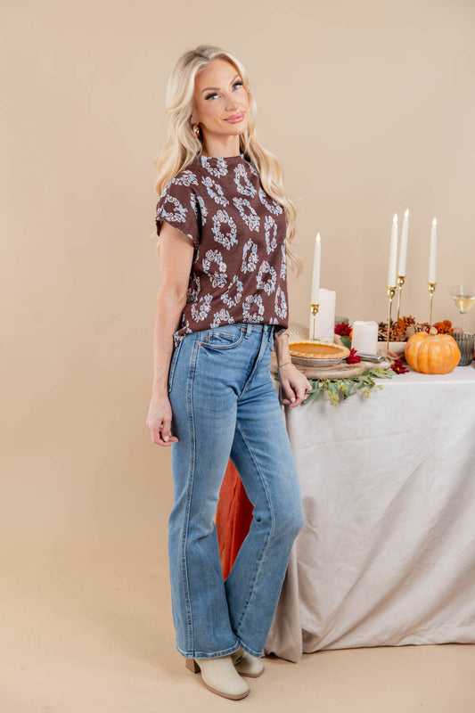 A woman modeling a brown short-sleeve top with a white floral pattern and light blue high-waisted flare jeans, standing next to a decorated table with candles, a pumpkin, and a pie against a beige studio background.