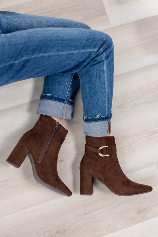A pair of brown suede ankle boots with block heels and a gold buckle detail, worn with cuffed blue jeans on a light wood floor.