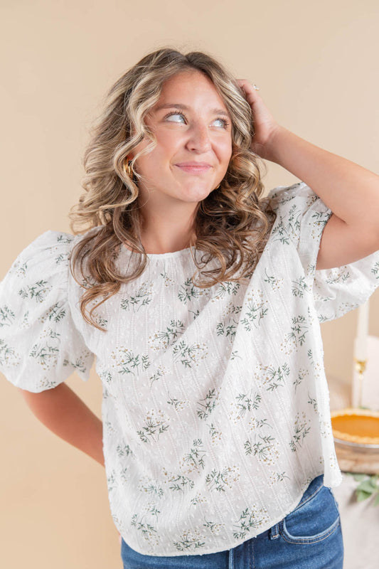 A woman wearing a white short-sleeve blouse with a delicate green and yellow floral pattern, paired with blue jeans, posing against a warm beige background.