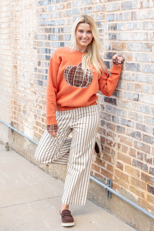 A woman posing against a brick wall wearing an orange crewneck sweatshirt with a plaid pumpkin graphic and cream pinstripe cropped pants.