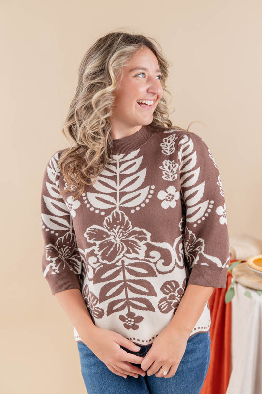 A woman wearing a brown and cream floral knit sweater with blue jeans, standing against a warm beige background.