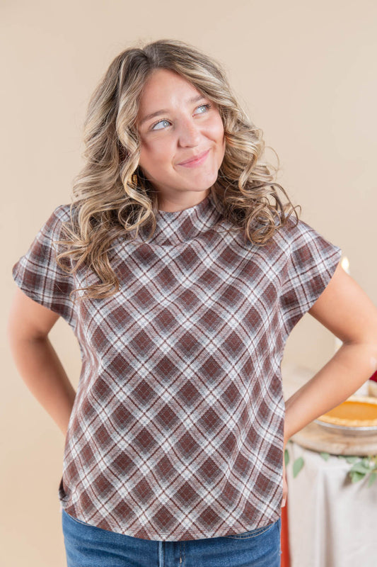 A woman with wavy blonde hair wearing a brown, beige, and white plaid short-sleeve mock neck top and blue jeans, posing with hands on hips against a warm beige background.