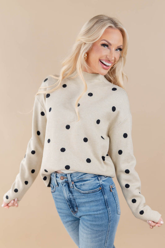 A smiling blonde woman wearing a cream knit sweater with black polka dots and light blue jeans, posed against a solid beige studio background.