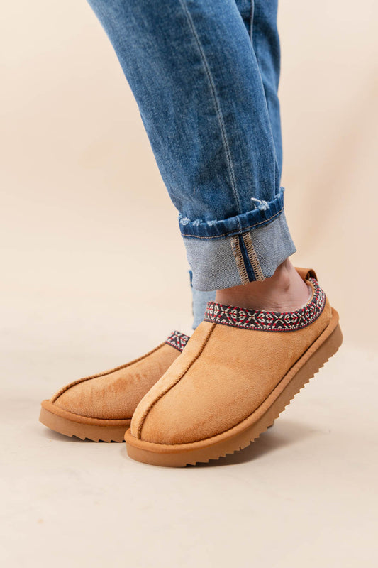 A woman wearing tan suede slip-on slippers with a patterned trim around the opening, paired with cuffed blue jeans, against a plain beige studio background.