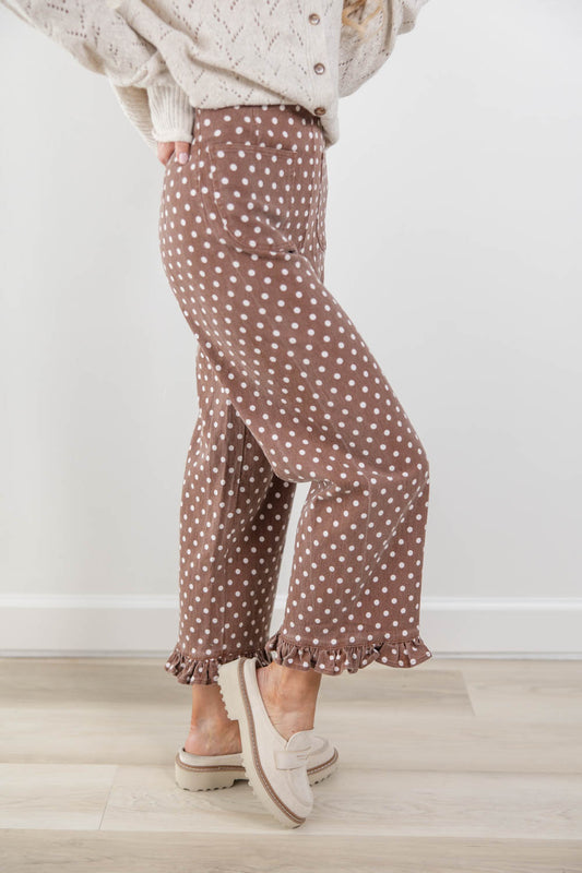 A woman wearing brown wide-leg pants with a white polka dot pattern and ruffled hems, styled with a cream knit sweater and cream slip-on shoes against a white wall and light wood floor.