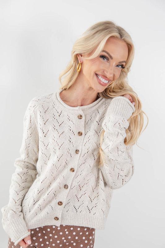 A smiling blonde woman wearing a cream pointelle cable-knit cardigan with brown buttons, paired with brown polka-dot pants, against a light gray studio background.